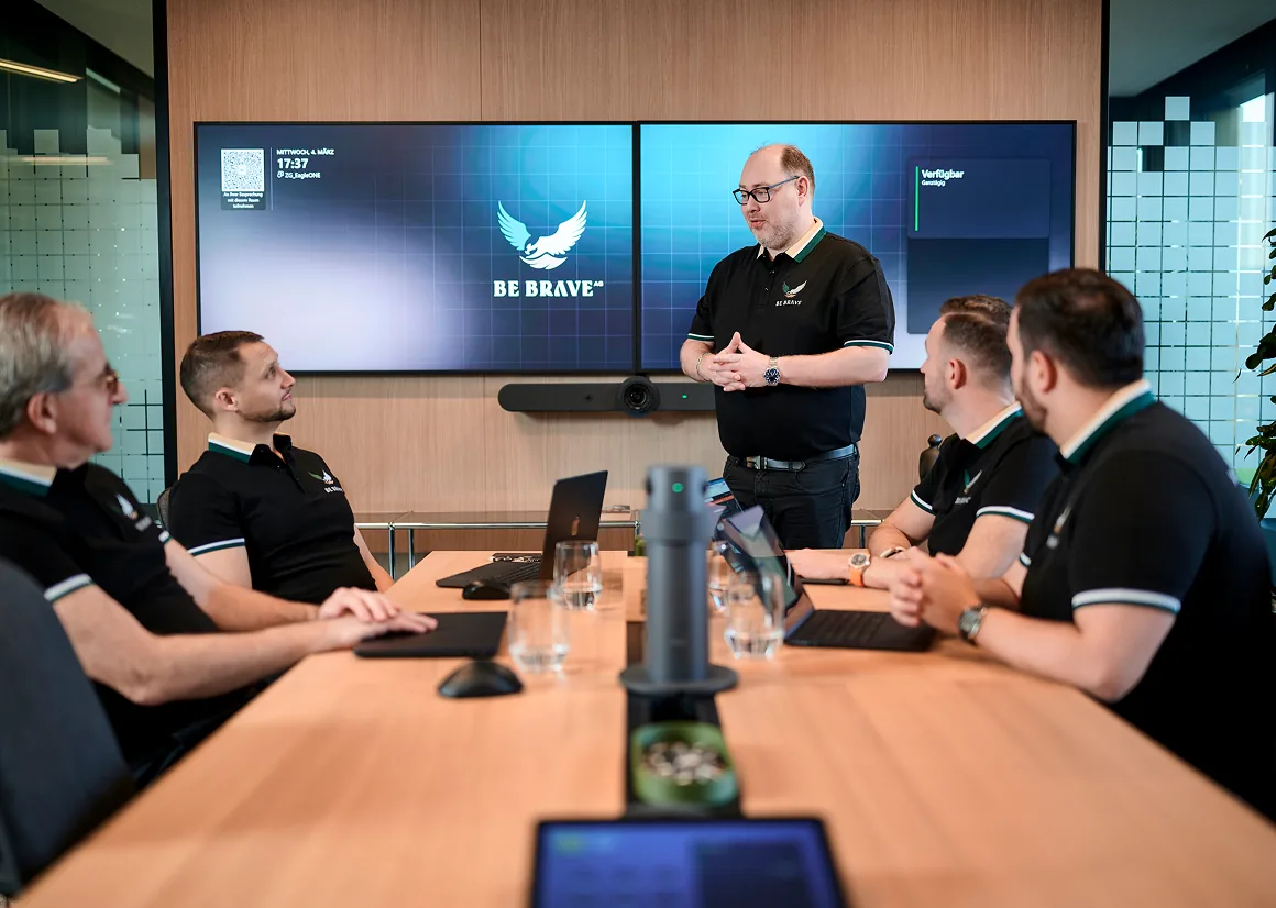 Besprechung im Konferenzraum: Ein Mitarbeiter präsentiert vor zwei Bildschirmen mit dem BE-BRAVE-Logo
