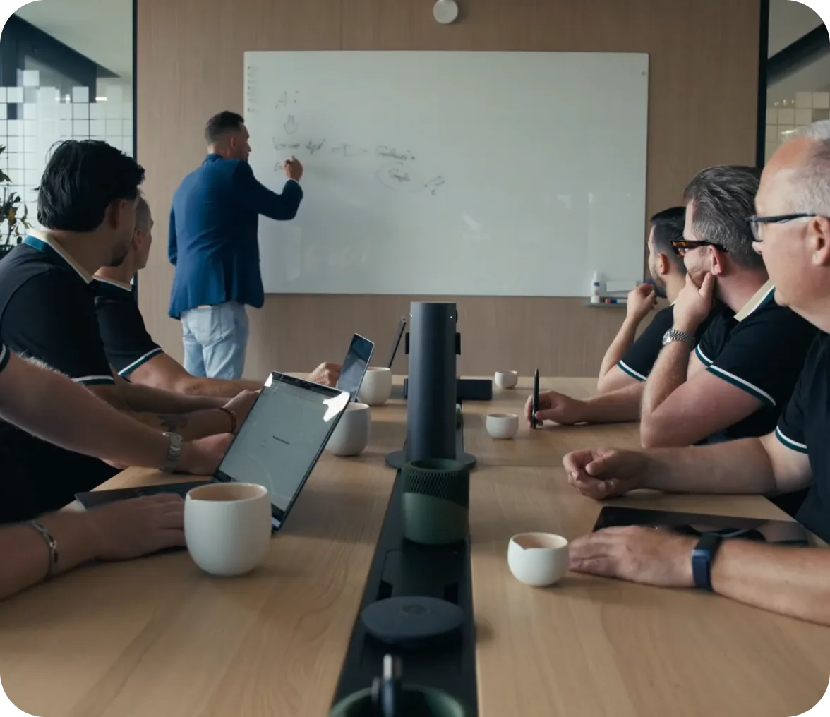Besprechung am Tisch mit Laptops, im Hintergrund schreibt ein Mann an ein Whiteboard